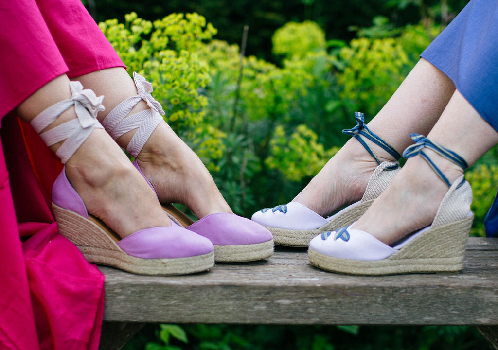 Two pairs of women's wedge sandals in lilac and blue on a wooden bench with a green outdoor background.