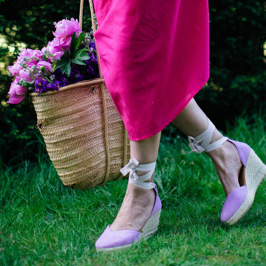 Person wearing purple shoes with white bows, standing on grass with a basket of flowers.