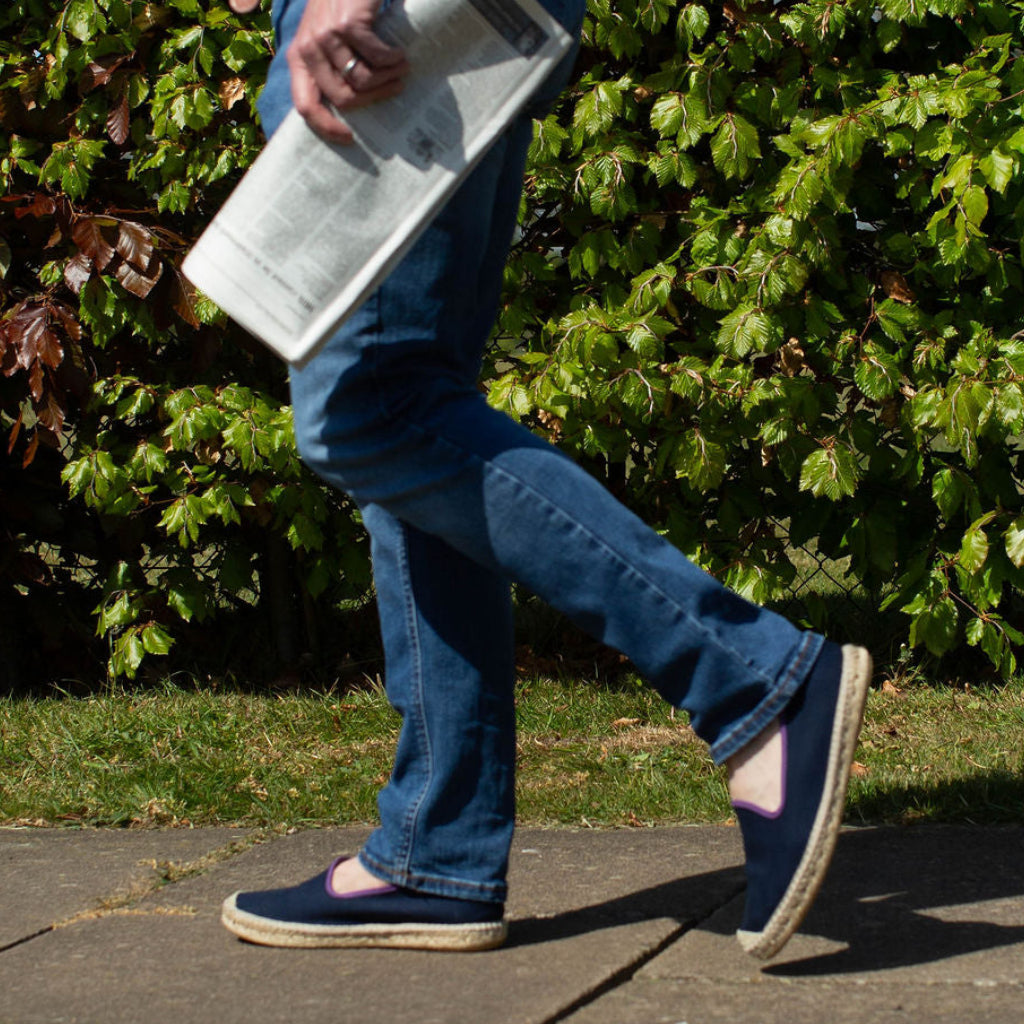 Person walking outdoors holding a newspaper, with greenery in the background wearing denim jeans and blue loafers.