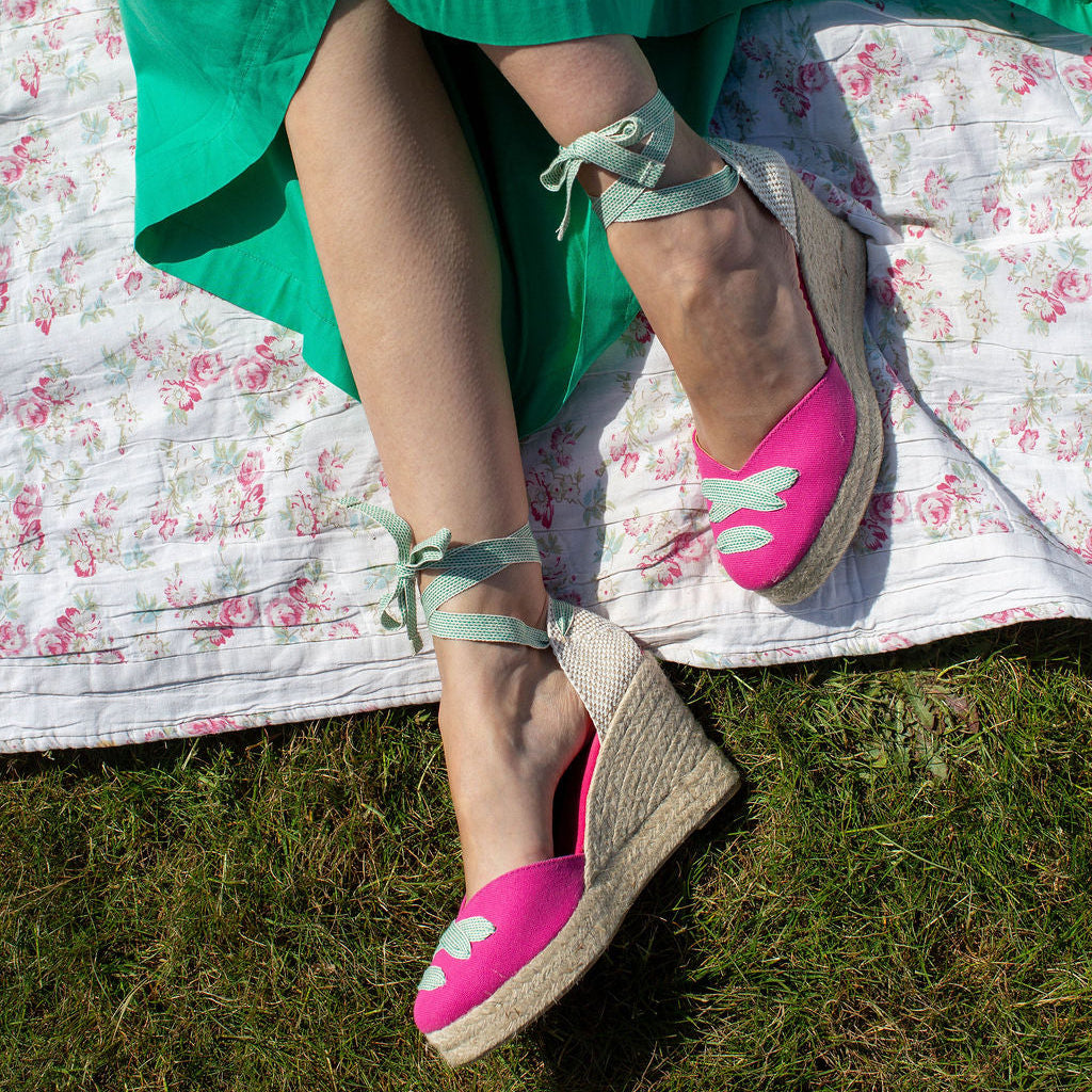 Person wearing pink espadrilles with green fabric and floral blanket in the background