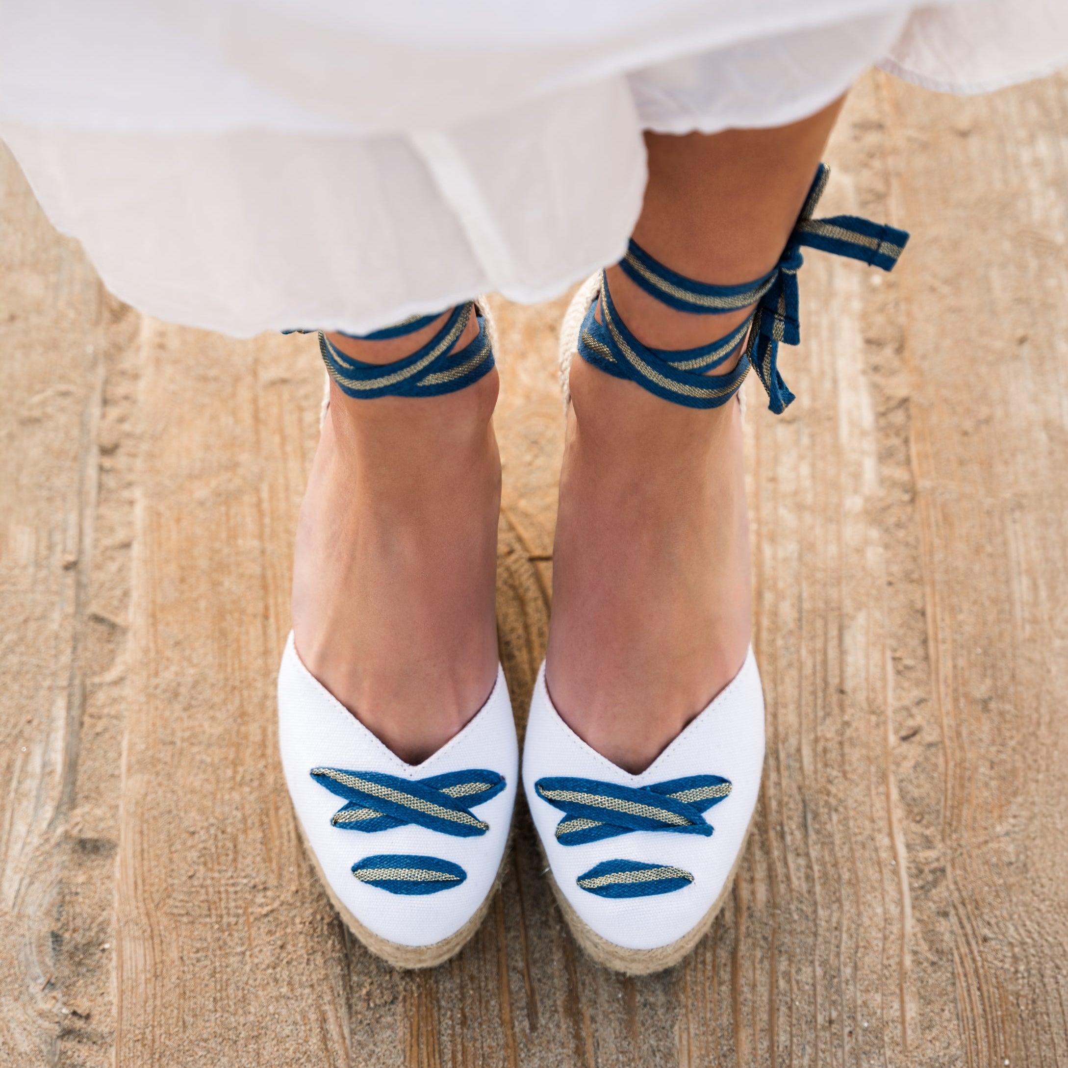 A women stands on a wooden sandy path. Only her feet and lower legs are visible. Wearing a long white skirt.  The white cotton wedge espadrilles have blue and gold ribbons tied in bows at her ankles and have the ribbon detailing across the almond shaped toe of the shoe. 