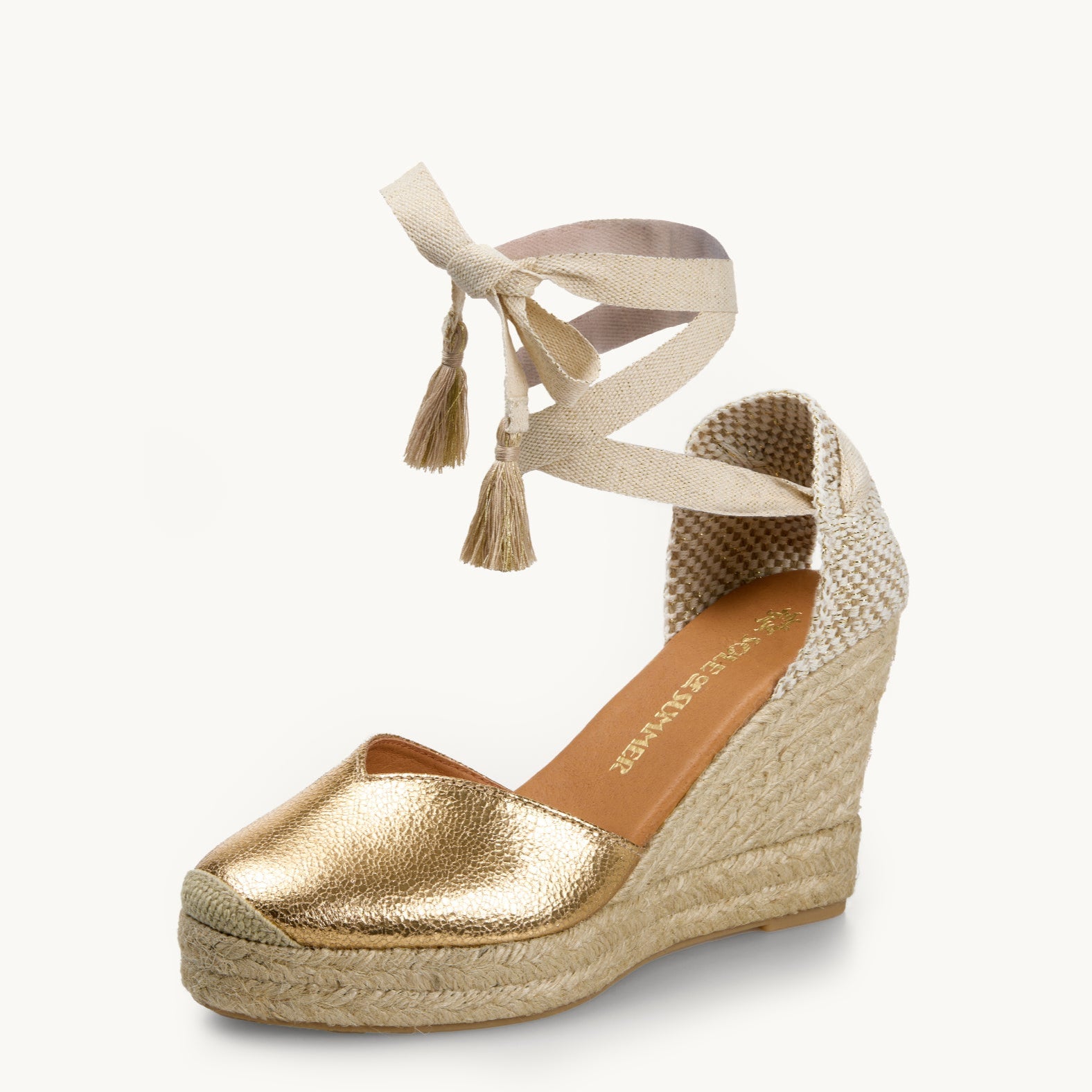 One crackled effect gold wedge espadrille displayed with white ribbons with gold tassels on the ends tied in a bow as if around an ankle.  The sole is made of Jute. It has an almond shaped toe. The insole is lined in tan leather with the gold sole of summer logo showing.