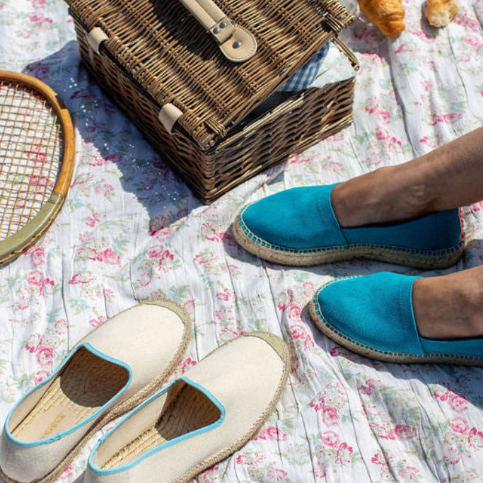 Picnic scene with vintage tennis racket, wicker picnic basket, and turquoise shoes on a floral blanket.