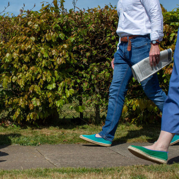 Two people walking along a path with greenery behind, one carrying a newspaper. Both wearing green shoes.