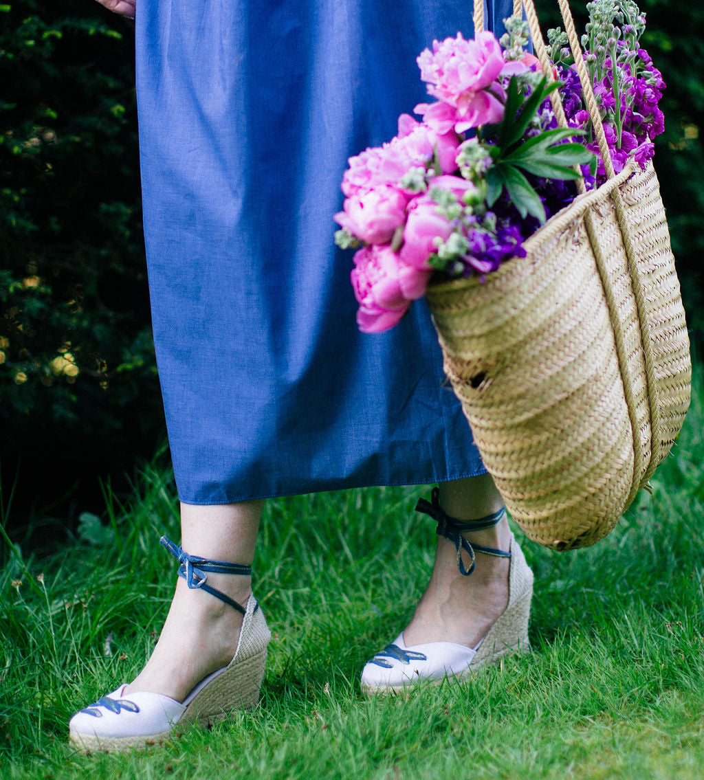 Person wearing a blue dress and white shoes with blue accents, holding a straw bag filled with flowers on grass.